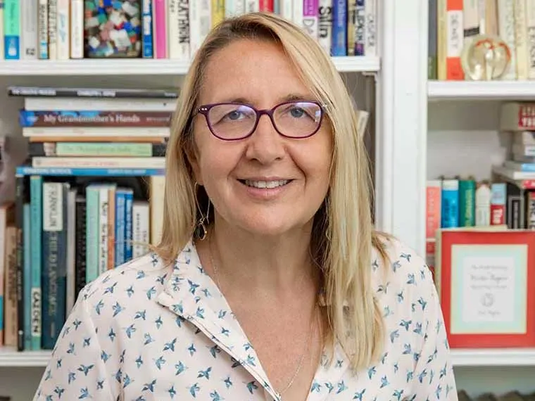 Person with glasses and long hair smiling, standing in front of a bookshelf filled with books and framed pictures.