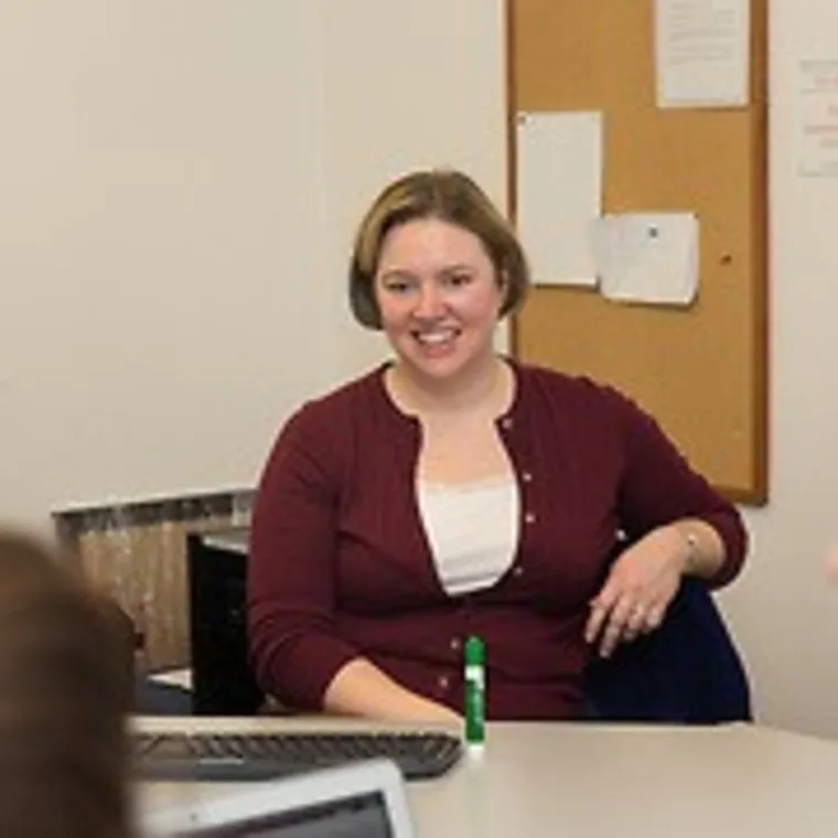 Person sitting at a desk, smiling, with a bulletin board in the background. A laptop and pen are on the desk.