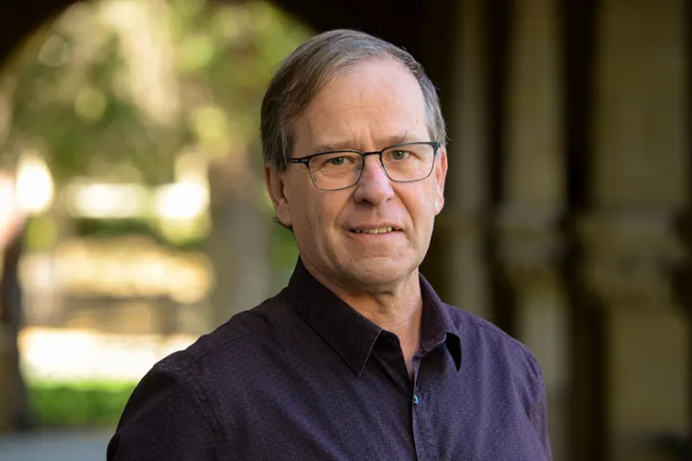 Person with glasses and short hair wearing a dark shirt, standing in an outdoor setting with blurred trees and arches in the background.
