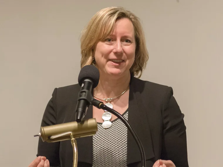 Person speaking at a podium with a microphone, wearing a black blazer and patterned shirt, smiling slightly. Neutral background.