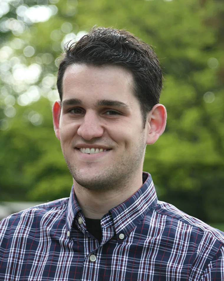 Person smiling, wearing a plaid shirt, with short dark hair. The background shows blurred green foliage.