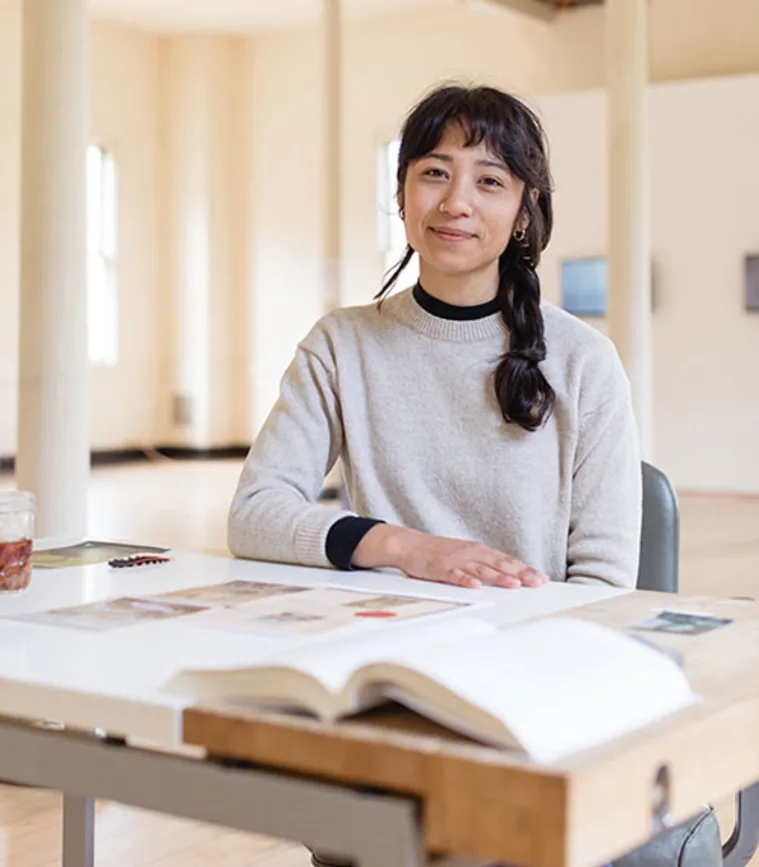 Person sitting at a table with books and papers, in a bright room with high ceilings and columns, looking at the camera with a slight smile.