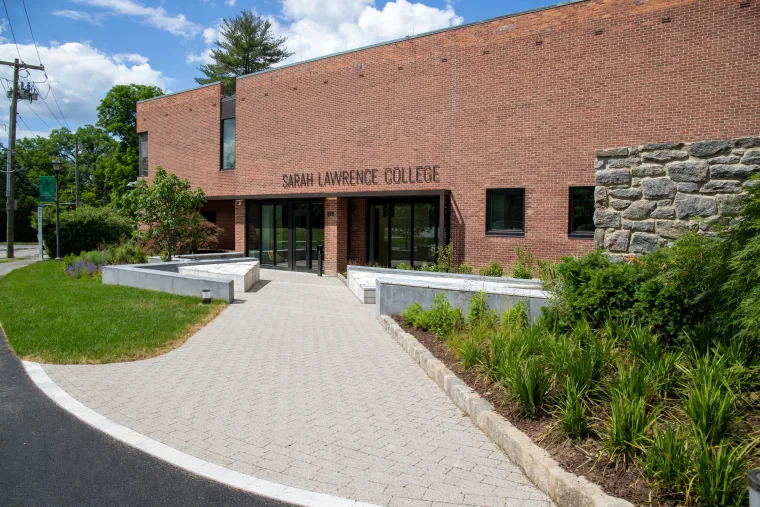 Brick building with "Sarah Lawrence College" on the facade, surrounded by trees and greenery under a blue sky.