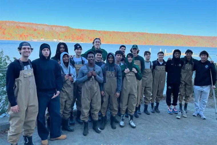 Group of people wearing waders and hoodies pose by a riverside with autumn foliage in the background.
