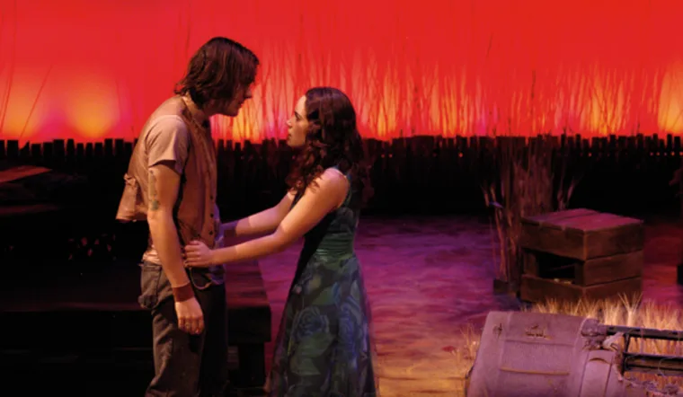 Two people stand closely on a stage with a dramatic red and orange backdrop, surrounded by props like crates and tall grass.