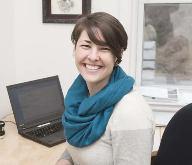 Person sitting at a desk with a laptop, smiling, wearing a teal scarf. A window and framed picture are visible in the background.