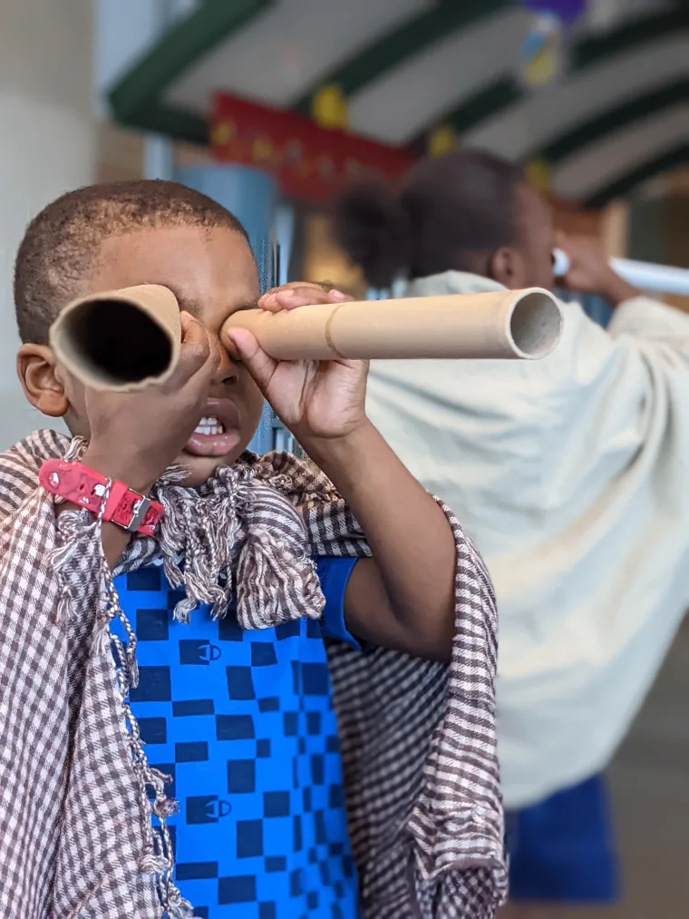 Two children playfully look through cardboard tubes, wearing capes. One is in focus, smiling, with a red watch and checkered shirt.