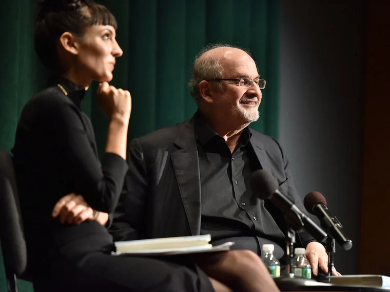 Two people seated on a stage, one with a notebook, the other smiling with microphones in front. Green curtain backdrop.
