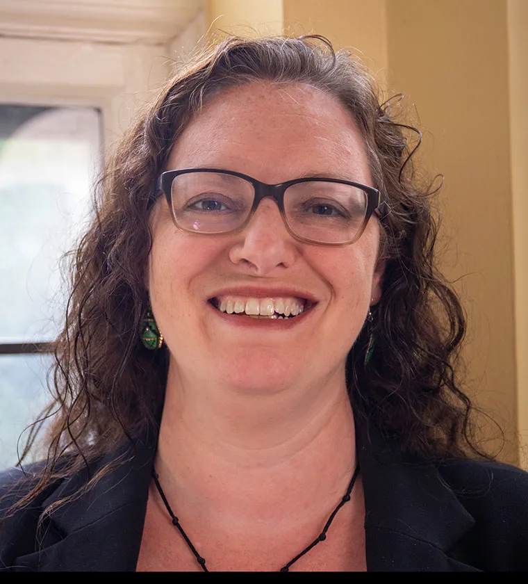 Person with wavy hair and glasses smiling in a warmly lit room, wearing earrings and a dark jacket.
