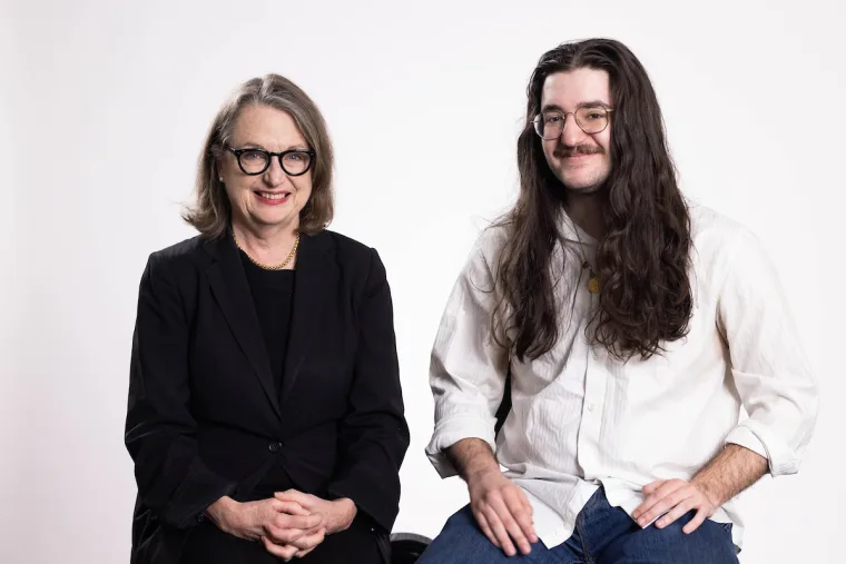 Two people sit side by side, smiling. One wears a black suit and glasses, the other has long hair and a white shirt with glasses.
