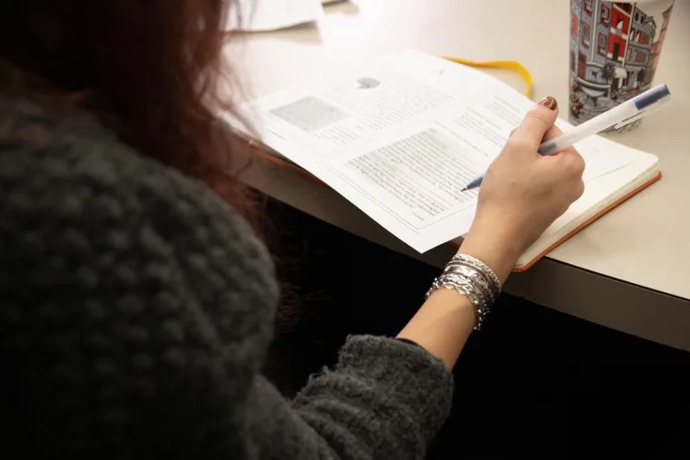 Person in grey sweater writing notes in an open book on a desk, with a patterned coffee cup nearby.