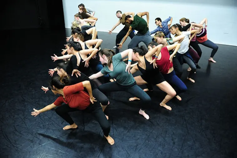 A group of dancers in colorful shirts and black pants performing a synchronized, dynamic pose on a stage with a dark floor.