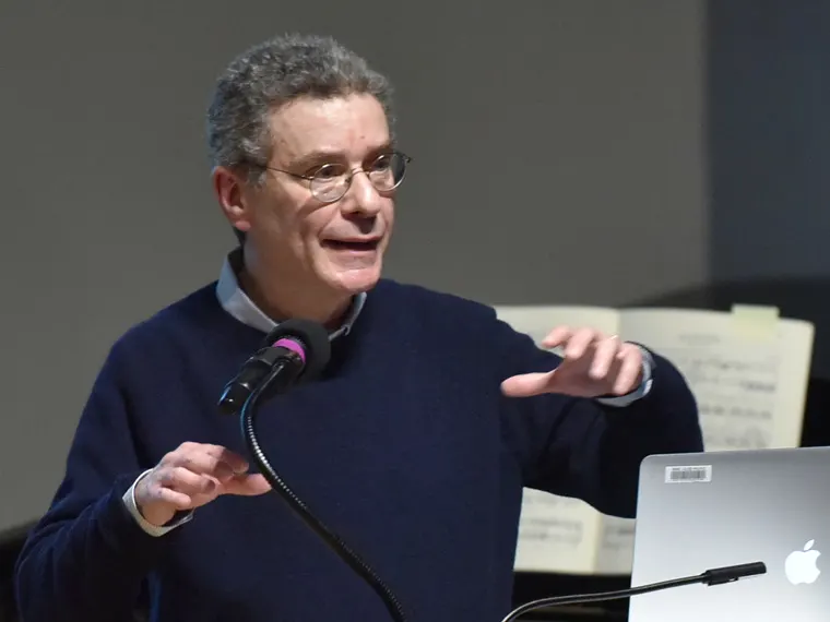 Person speaking at a microphone with a laptop in front, gesturing with one hand. Sheets of paper are visible in the background.