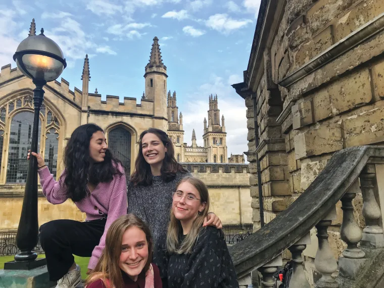 Four people smiling and posing near a historic building with spires, under a cloudy sky. One is sitting on a stone ledge.