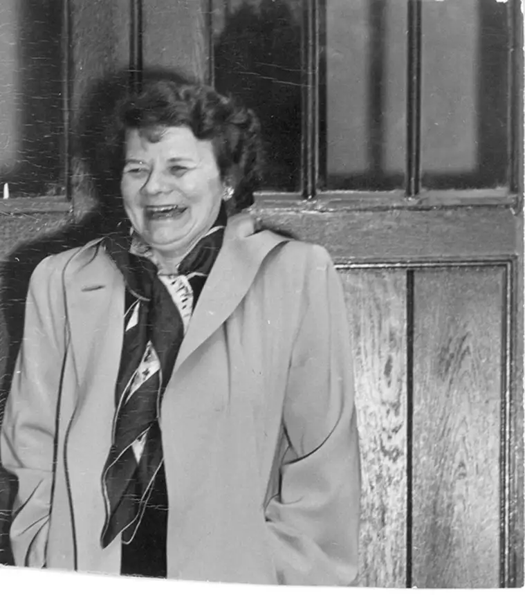 Person in a light coat and scarf smiling in front of a paneled wooden door.
