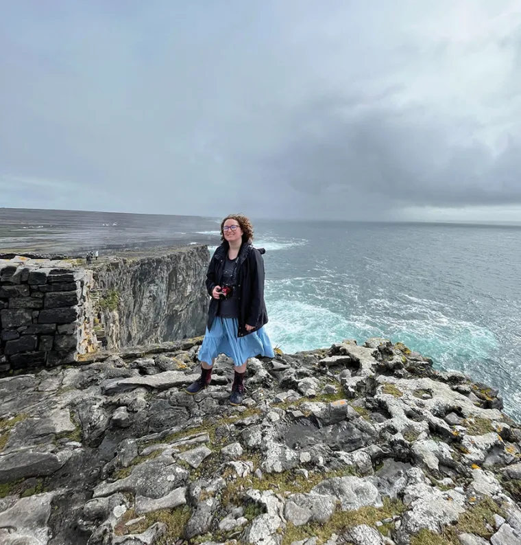 Person with curly hair stands on rocky cliff holding a camera, overlooking the ocean with cloudy sky in the background.
