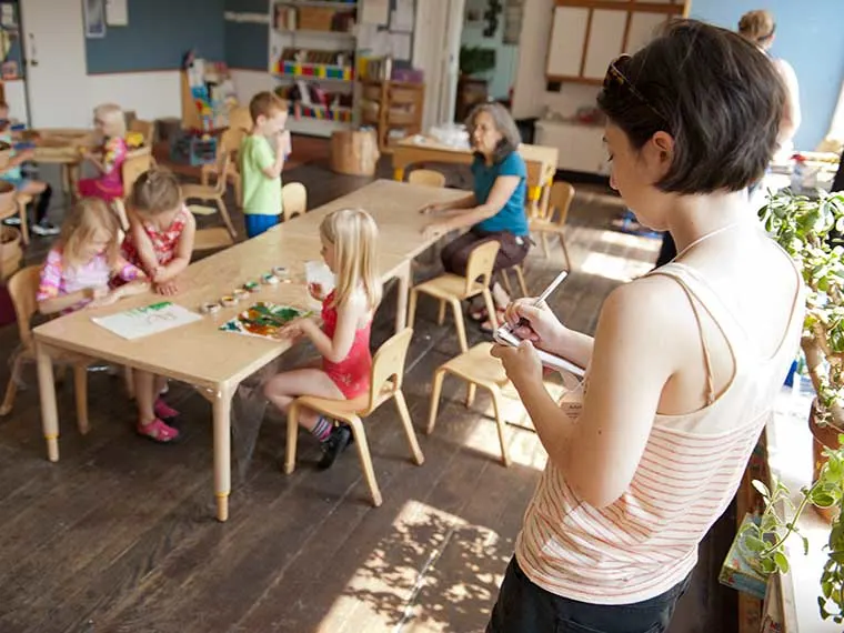 Classroom scene with children sitting at a table doing crafts, an individual writing on a notepad nearby, and an adult sitting in the background.