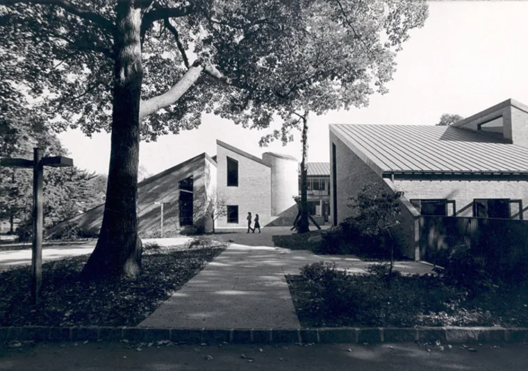 Campus sidewalk lined with trees and modern buildings; two individuals walk along the path.