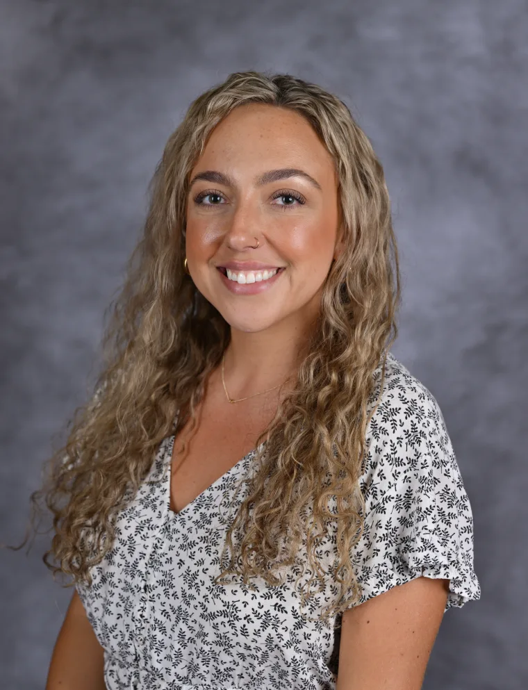 Person with curly hair smiling, wearing a patterned top against a gray background.