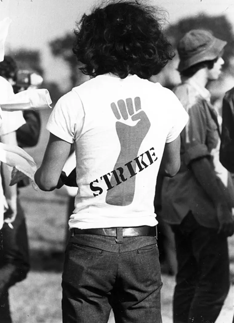 Person with curly hair wearing a shirt featuring a raised fist and the word "STRIKE" in a group setting outdoors.