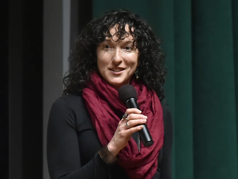 Person with curly hair speaking into a microphone, wearing a maroon scarf and black outfit, standing in front of a green curtain.