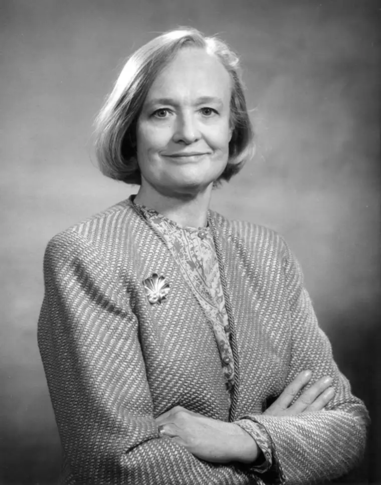 Person with short hair wearing a patterned jacket and brooch, arms crossed, smiles slightly in a black-and-white portrait.