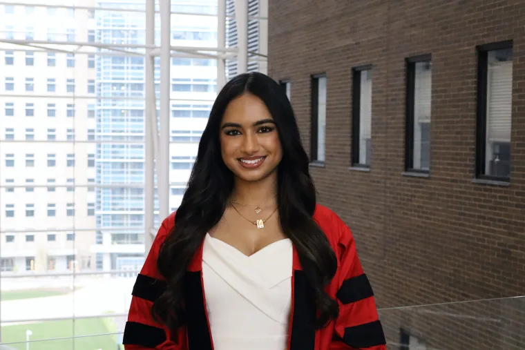 Person in a white outfit and red-striped robe smiles indoors, with a cityscape visible through large windows in the background.