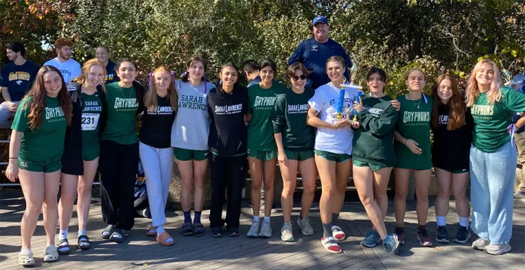 A group of seventeen people wearing "Sarah Lawrence" and "Gryphons" shirts, standing outdoors on a sunny day, some holding a trophy.