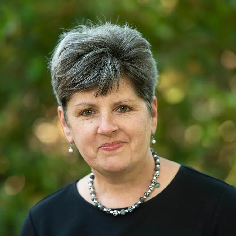 Person with short hair and a necklace, in front of a blurred green background.