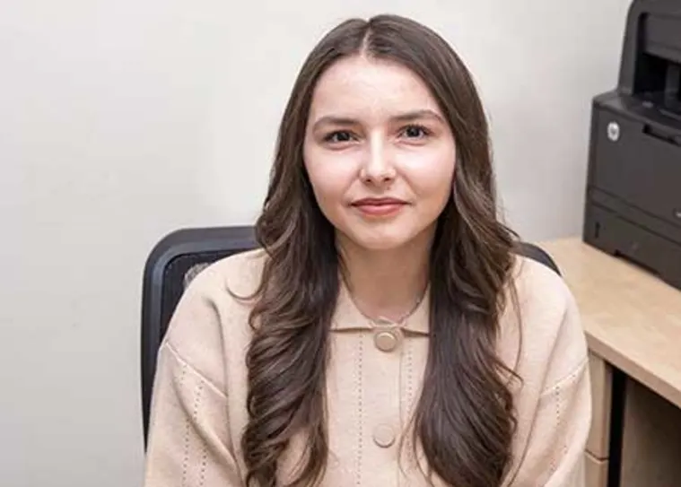 Person with long hair sits in an office chair, wearing a beige sweater. A printer is seen on a desk in the background.