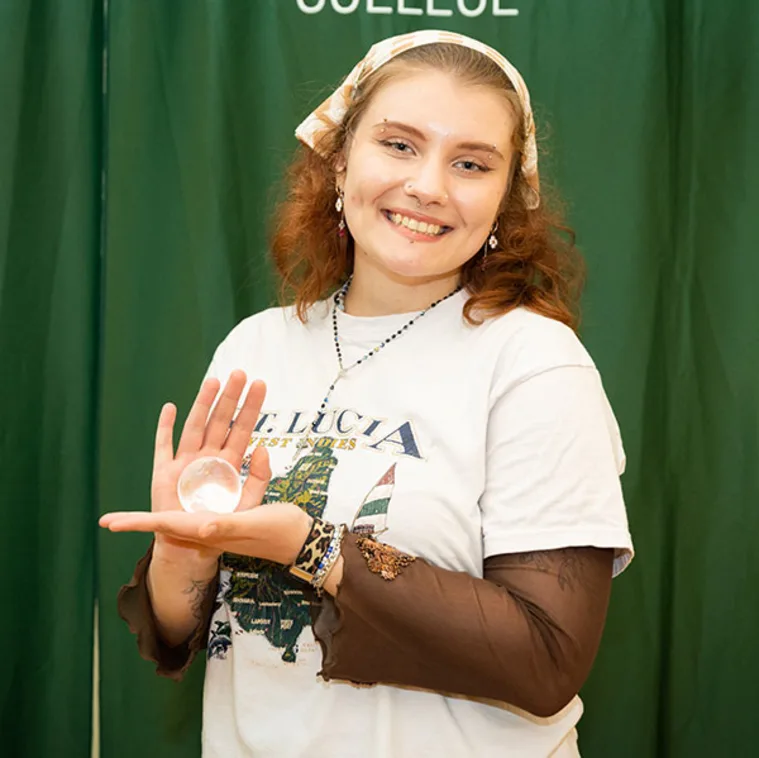 Person smiling, holding a crystal ball in one hand, stands in front of a green background with white text. Wearing a T-shirt and patterned headscarf.