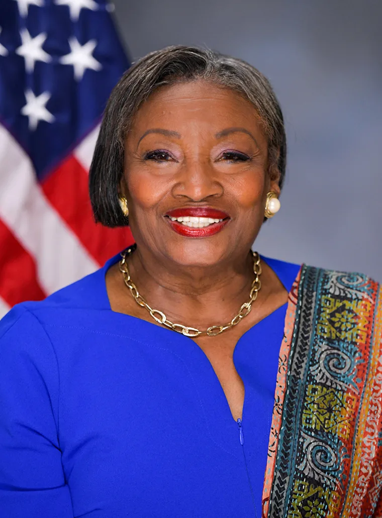 Person smiling, wearing a blue top and patterned shawl, with short hair. An American flag is partially visible in the background.