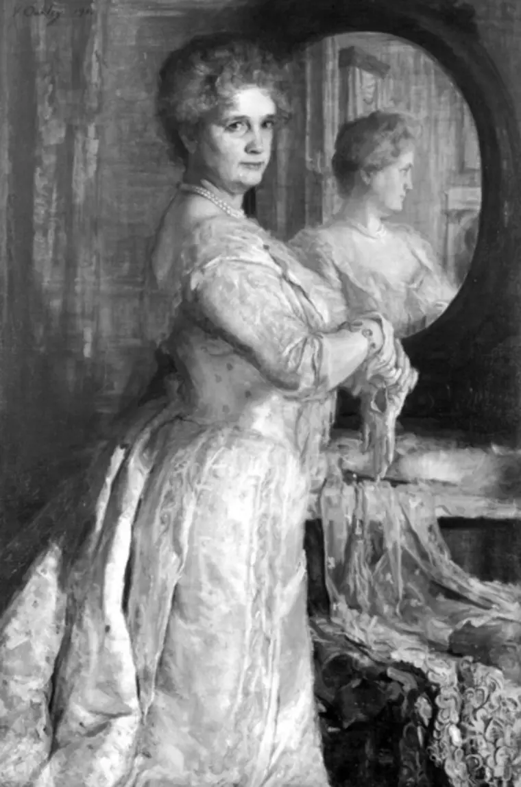 Person in an ornate dress standing by a vanity mirror, looking over with a direct gaze, reflection visible. The setting is elegant and classical.