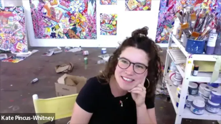 Person with curly hair and glasses smiles in an art studio. Colorful paintings and art supplies are visible in the background.