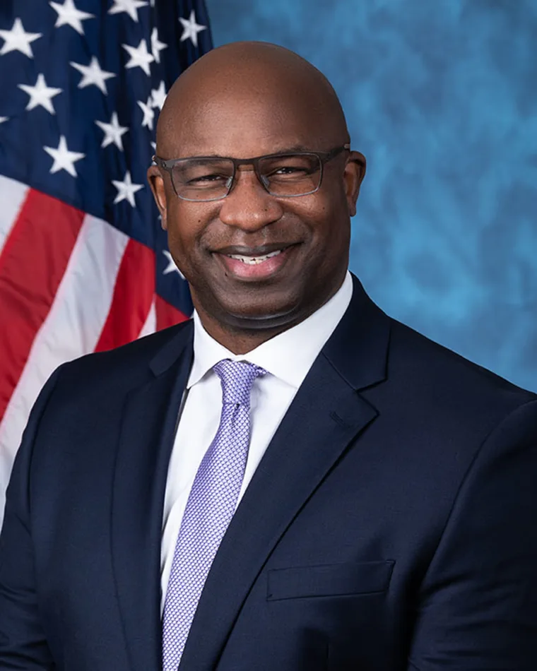 Person in a suit with glasses smiling in front of an American flag and a blue background.