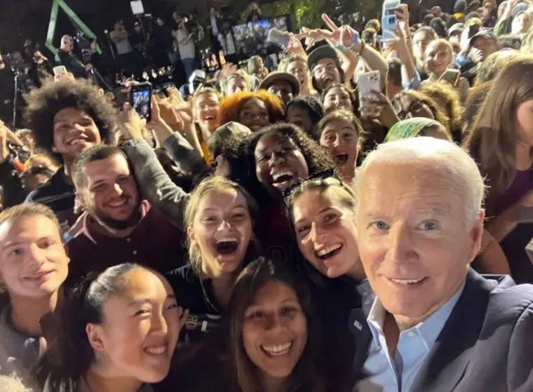 Group of smiling people taking a selfie outdoors, with a prominent individual in the foreground and a crowd in the background holding up phones.