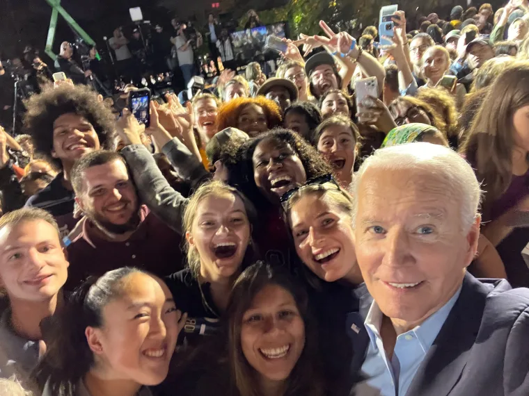 A group of smiling individuals taking a selfie together at an outdoor event with a large, cheerful crowd in the background.