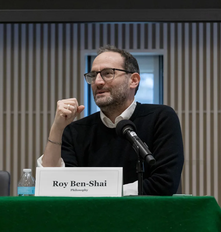 Person with glasses speaks into a microphone at a panel. Nameplate reads "Roy Ben-Shai, Philosophy." Background has vertical wooden slats.