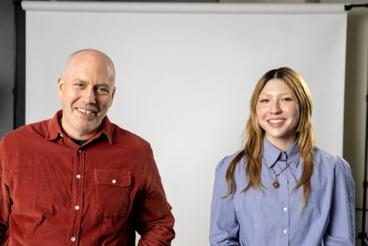 Two people smiling, standing in front of a white backdrop. One is wearing a red shirt, the other a blue shirt.