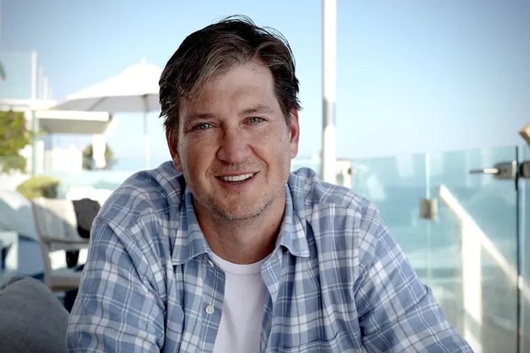 Person smiling at a sunny outdoor setting with ocean view, wearing a blue plaid shirt, with white umbrellas and glass railings in the background.
