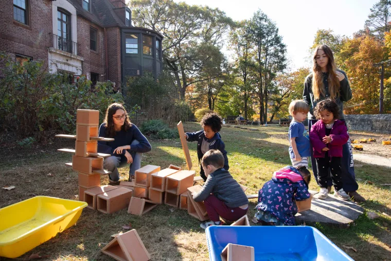 People and children building with large blocks outdoors near a brick building.