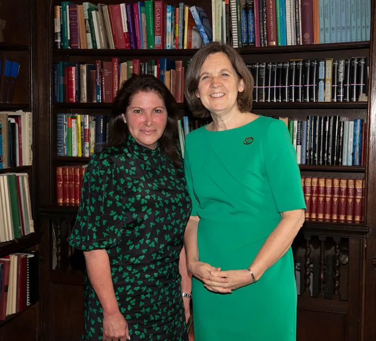 Two people smiling in front of a filled bookshelf. One wears a green dress, the other a dark dress with green patterns.