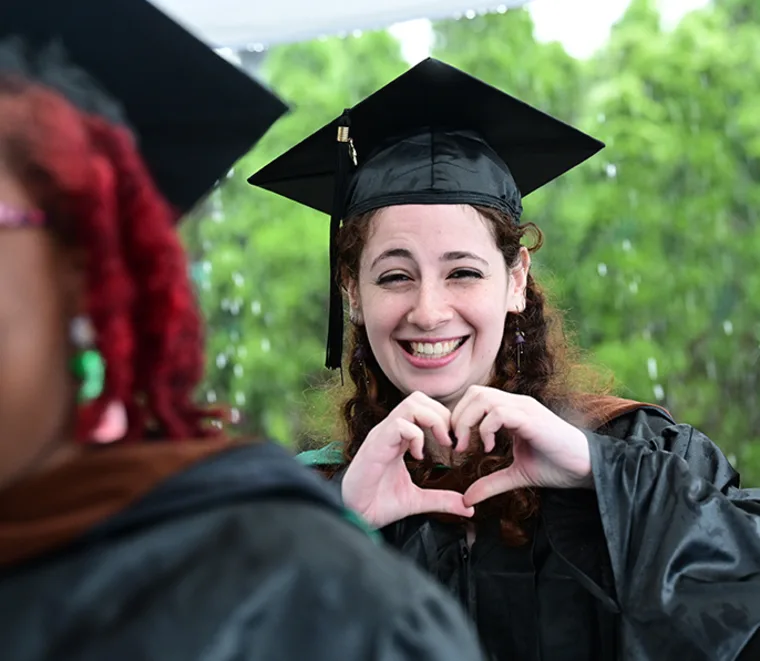 Graduate in cap and gown smiling, forming a heart shape with hands. Another graduate is partially visible in the foreground.