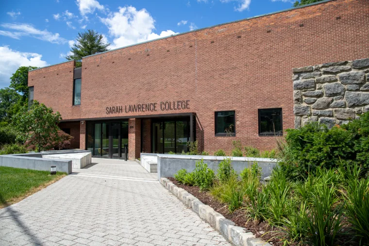 Sarah Lawrence College building with a brick facade, stone wall section, and pathway surrounded by greenery under a blue sky.