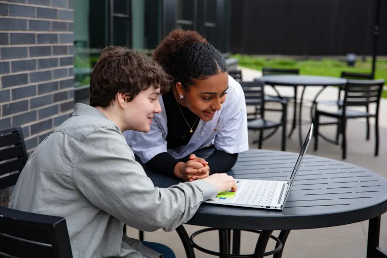 Two people sitting at a round outdoor table, looking at a laptop screen and smiling, with chairs and a patio visible in the background.