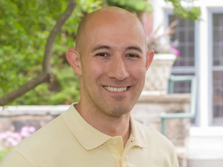 Person smiling outdoors, wearing a light yellow collared shirt, with blurred greenery and building in the background.