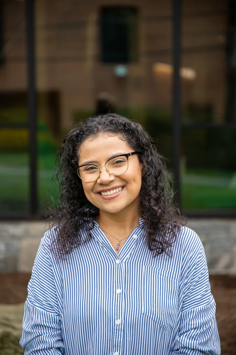 Person with curly hair and glasses smiling, wearing a blue and white striped shirt, standing in front of a window.