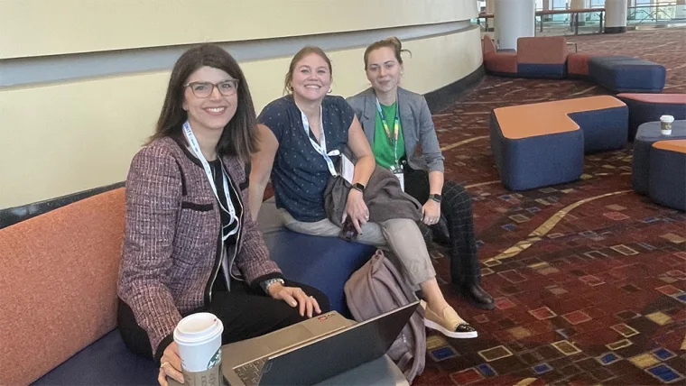 Three people sitting together on a bench in a conference area, one holding a laptop and another holding a coffee cup, smiling at the camera.
