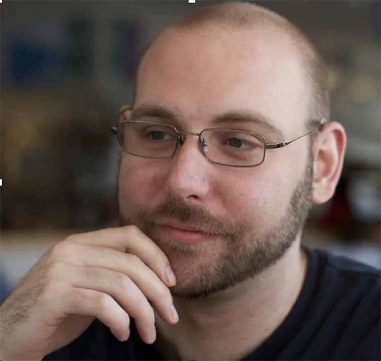 Person with glasses and beard, looking thoughtful with hand on chin, in a softly blurred indoor setting.