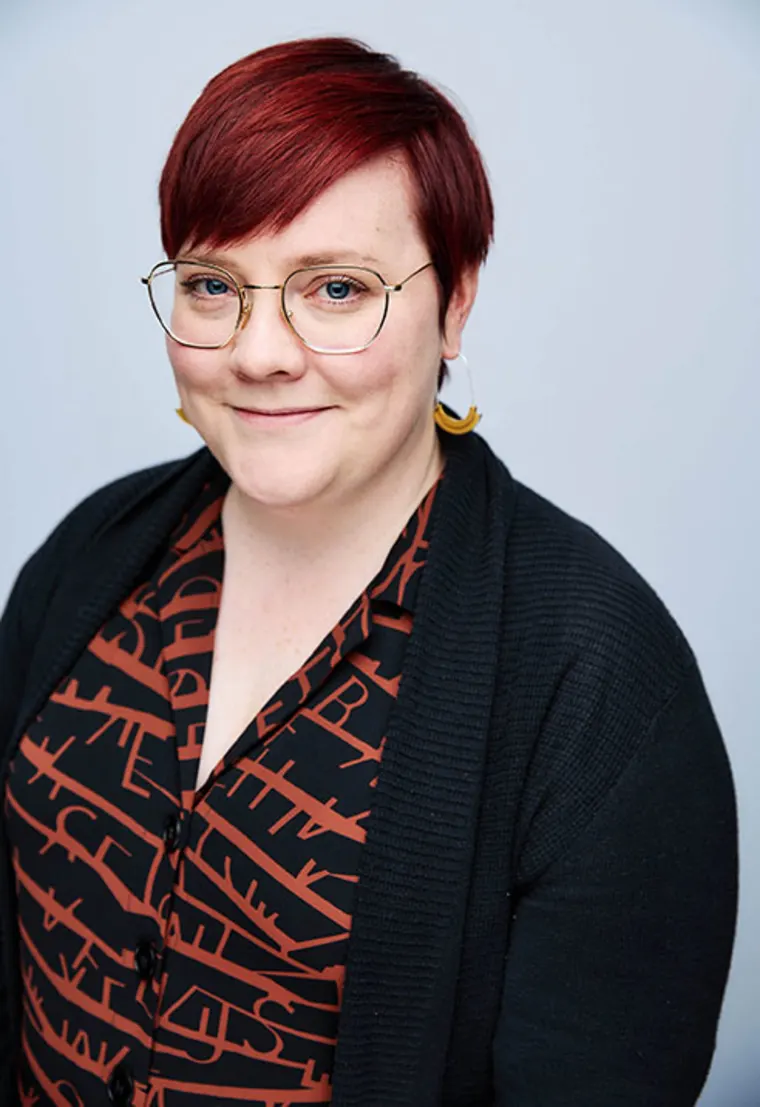 Person with short red hair, wearing glasses, patterned shirt, and dark cardigan, smiling at the camera against a light background.